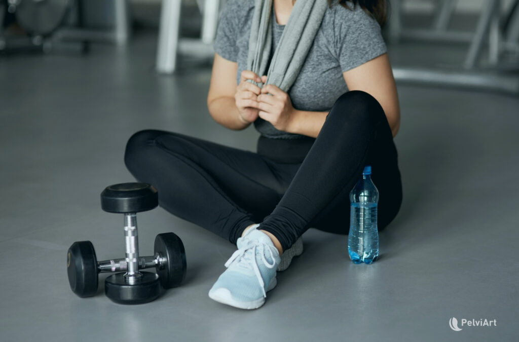 Mujer deportista sentada en el gimnasio con una toalla, una mancuerna y una botella de agua, en pausa durante su entrenamiento